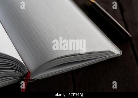 Minimalistic close-up of an open blank notebook with grid pages and a red bookmark on a dark wooden table. A pen is slightly blurred in the background Stock Photo