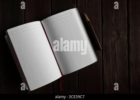 Top view of an open empty notebook with grid paper and a pen lying on a dark wooden surface. Perfect for themes of journaling, education, planning, or Stock Photo