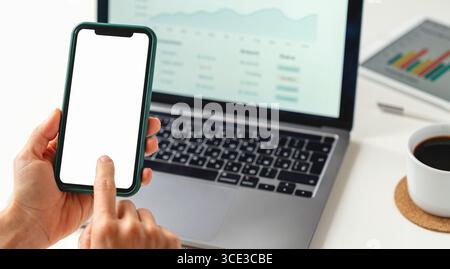 Digital banking and mobile technology. Hands holding smartphone with blank screen against laptop with financial interface Stock Photo