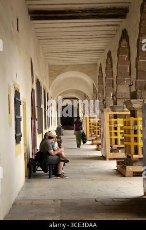 A view of a vaulted stone corridor in a historic building, with tourists resting on a bench and others walking further down the passage. Stock Photo