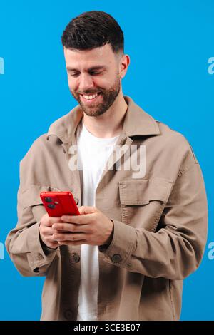 Man smiling while using a red smartphone against a bright blue background Stock Photo