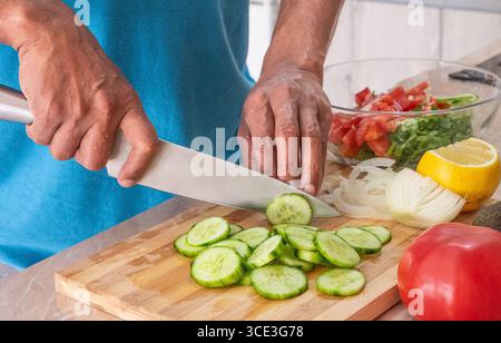 Close Up Photo of man Hands Making Fresh cucumbers on a Table Full with Organic Vegetables. Hands, cutting board and salad in kitchen with green veget Stock Photo