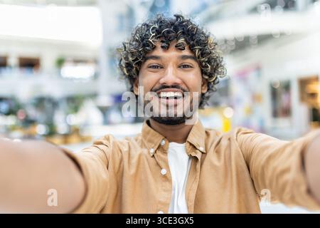 A joyful young man smiling confidently while taking a selfie in a contemporary indoor setting, showcasing happiness and self-confidence. Stock Photo