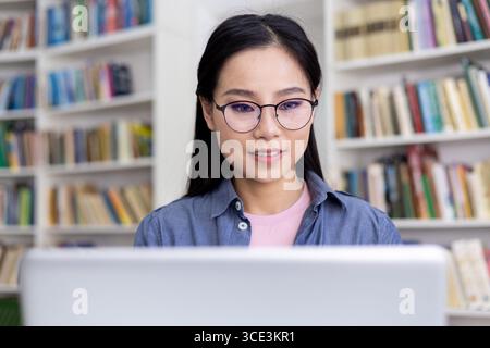 A young woman wearing glasses working on a laptop in a library setting. Shelves filled with books are arranged in the background, creating a calm and intellectual atmosphere while she concentrates. Stock Photo