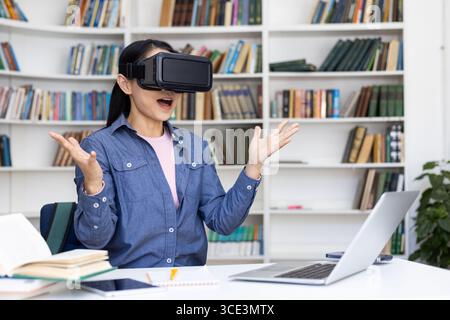 A woman explores virtual reality using a VR headset in a library, displaying excitement and amazement. She is engaging with innovative technology surrounded by books and working on her laptop. Stock Photo
