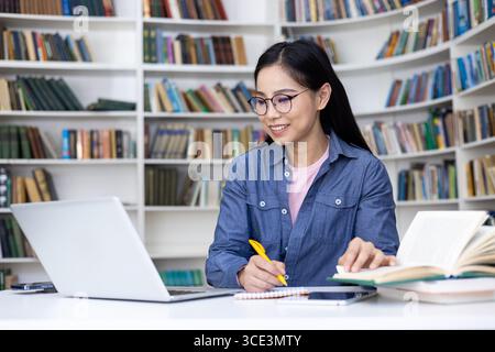 A young woman studies in a library, working on a laptop and taking notes, surrounded by shelves of books. Stock Photo