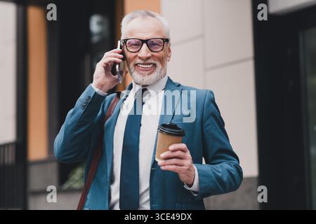 Successful businessman wearing a blue suit talking on a phone outdoors, holding coffee, with a confident smile Stock Photo