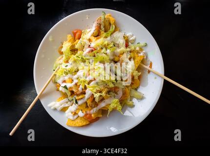 Top view of a plate of taco salad nachos topped with cheese, guacamole, shredded lettuce, and dressing, garnished with skewers on a dark background. m Stock Photo