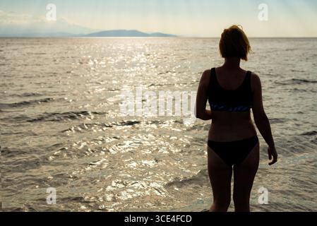 A woman standing in shallow water, wearing a black swimsuit, looking towards the shimmering sunset over the sea. Stock Photo