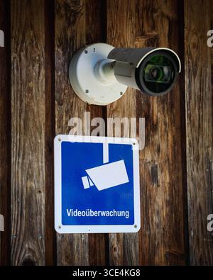 14.08.2025, Handenberg, AUT, Unterwegs in Oberösterreich, Themenbild, Symbolbild, Verschiedene Themenbilder, Videoüberwachung, Kamera, Überwachung, im Bild Das Bild zeigt eine Überwachungskamera, die an einer Holzoberfläche montiert ist. Die Kamera hat ein weißes Gehäuse mit einem runden Objektiv und ist durch Schrauben befestigt. Unter der Kamera hängt ein quadratisches Schild mit einem blauen Hintergrund und einem stilisierten Bild einer Kamera in Weiß. Darunter steht in klarer Schrift Videoüberwachung .Die Holzoberfläche wirkt rustikal und trägt zur Atmosphäre des Ortes bei. Die Kamera und Stock Photo