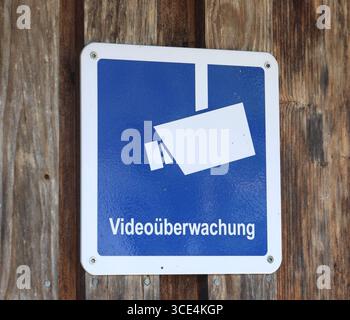 14.08.2025, Handenberg, AUT, Unterwegs in Oberösterreich, Themenbild, Symbolbild, Verschiedene Themenbilder, Videoüberwachung, Kamera, Überwachung, im Bild Das Bild zeigt eine Überwachungskamera, die an einer Holzoberfläche montiert ist. Die Kamera hat ein weißes Gehäuse mit einem runden Objektiv und ist durch Schrauben befestigt. Unter der Kamera hängt ein quadratisches Schild mit einem blauen Hintergrund und einem stilisierten Bild einer Kamera in Weiß. Darunter steht in klarer Schrift Videoüberwachung .Die Holzoberfläche wirkt rustikal und trägt zur Atmosphäre des Ortes bei. Die Kamera und Stock Photo