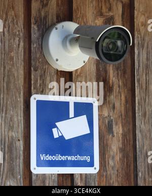 14.08.2025, Handenberg, AUT, Unterwegs in Oberösterreich, Themenbild, Symbolbild, Verschiedene Themenbilder, Videoüberwachung, Kamera, Überwachung, im Bild Das Bild zeigt eine Überwachungskamera, die an einer Holzoberfläche montiert ist. Die Kamera hat ein weißes Gehäuse mit einem runden Objektiv und ist durch Schrauben befestigt. Unter der Kamera hängt ein quadratisches Schild mit einem blauen Hintergrund und einem stilisierten Bild einer Kamera in Weiß. Darunter steht in klarer Schrift Videoüberwachung .Die Holzoberfläche wirkt rustikal und trägt zur Atmosphäre des Ortes bei. Die Kamera und Stock Photo