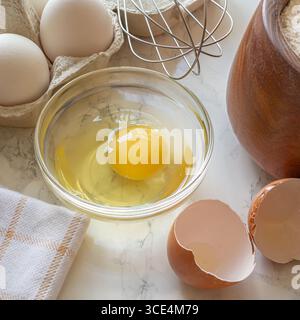 Fresh eggs are placed in a bowl and on a countertop alongside a whisk, flour, and eggshells, ready for baking. Stock Photo