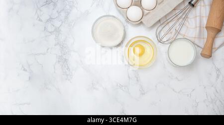 Ingredients for baking are arranged on a marble countertop with eggs, flour, and milk ready for mixing and cooking. Stock Photo