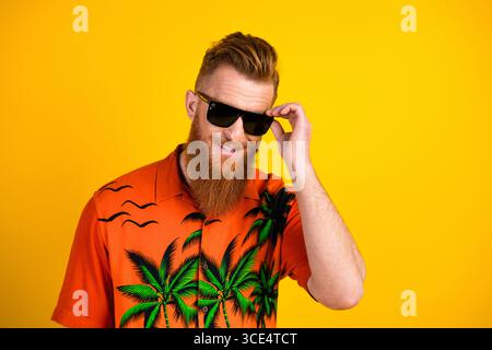 Cheerful young man with red beard dressed in vibrant tropical shirt poses confidently against yellow backdrop Stock Photo