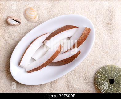 Fresh coconut meat, in oval white bowl, on beach sand with shells. Opaque white flesh of a fresh and young coconut, cut into strips, and ready to eat. Stock Photo
