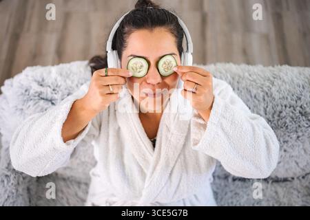 Woman applying cucumber slices on eyes while listening to music and wearing bathrobe Stock Photo