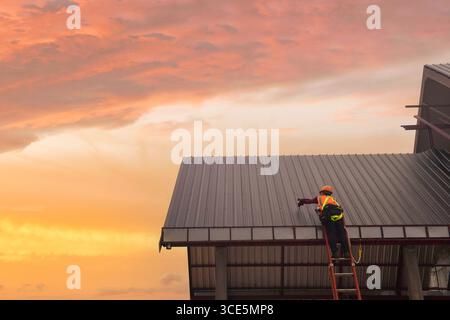 Rear view of a roofer standing on a ladder attaching metal sheets on a roof, Thailand Stock Photo