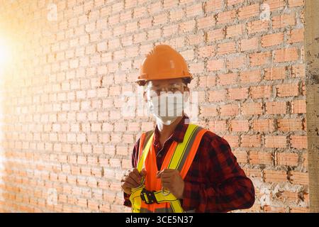 Portrait of a construction worker standing in front of a brick wall wearing a protective face mask, hard hat and reflective clothing, Thailand Stock Photo