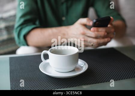 Close-up of a man sitting at a table using his phone while drinking a cup of black coffee Stock Photo