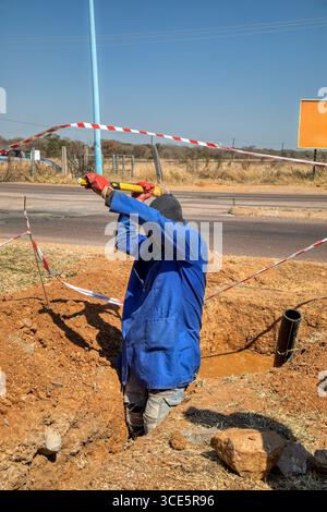 Single African road construction worker with a pickaxe digging a trench ditch on the side of the road, infrastructure repairs Stock Photo
