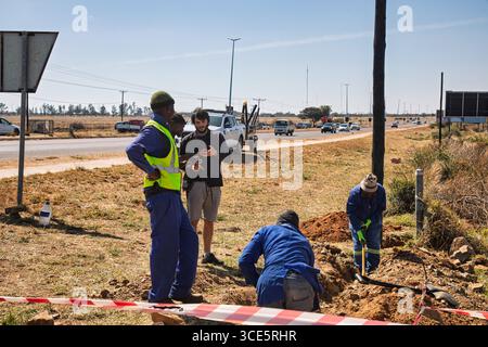 African road construction workers with a pickaxe and shovel , manager showing a plan on the phone ,digging a trench ditch on the side of the road, inf Stock Photo