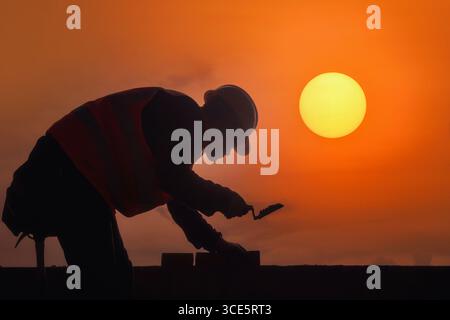 Side view silhouette of a bricklayer laying bricks on a construction site at sunset, Thailand Stock Photo