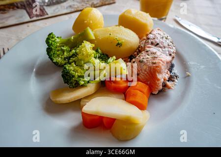 A healthy dinner of baked salmon with boiled potatoes and vegetables served on a white plate on a dining table. Stock Photo