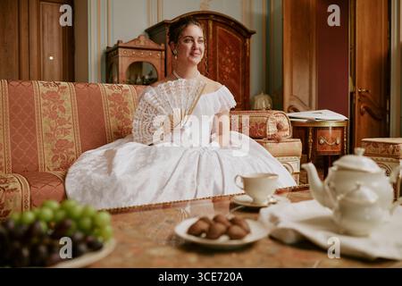 Caucasian young adult woman sitting on ornate sofa holding lace fan wearing historical dress, smiling slightly with hair styled up table with tea set and fruit in foreground Stock Photo