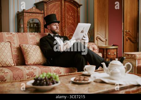 Caucasian middle aged man with beard wearing formal suit and top hat sitting on ornate sofa reading newspaper, holding paper with gloved hands, tea set and fruit on table Stock Photo