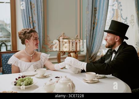 Caucasian young adult woman and Caucasian young adult man sitting at table dressed in formal historical attire holding hands and smiling during elegant tea gathering Stock Photo