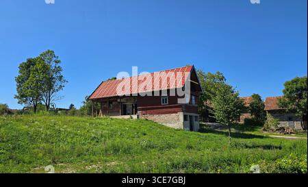 Traditional country barn with fresh red tiled roof and exposed brick sits on grassy field, surrounded by trees, nearby houses, and blue sky Stock Photo