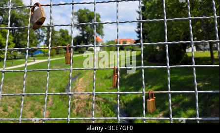 Multiple padlocks hang on grid-pattern fence, symbolizing love and commitment, with walking paths, trees, and lawns visible through the wire Stock Photo