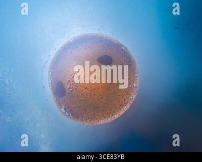 Coffee foam creating beautiful patterns inside glass cup seen from below, with blue background and condensation drops on the glass, perfect for backgr Stock Photo