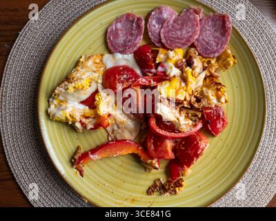 Scrambled eggs with chopped tomatoes, peppers and slices of salami are served on a yellow plate placed on a placemat, offering a simple yet satisfying Stock Photo