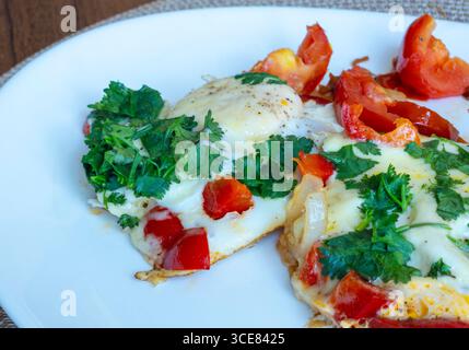 Close-up of fried eggs seasoned with black pepper and garnished with fresh parsley, accompanied by sliced tomatoes, onions, and bell peppers on a whit Stock Photo