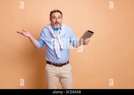 Confident mature businessman holding a tablet and shrugging against a beige background, showcasing stylish casual elegance Stock Photo