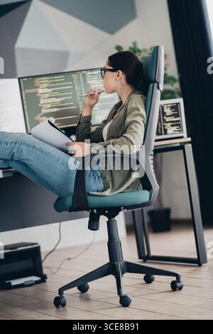 Young woman programmer analyzing code on dual monitors in a modern workspace with casual and professional ambiance Stock Photo