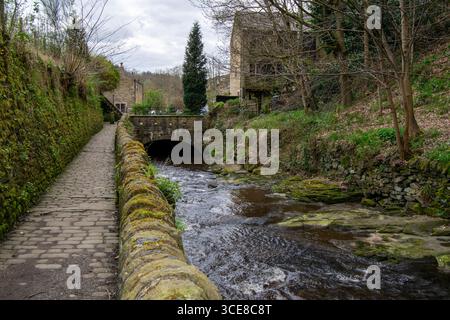 A stone-paved path and a mossy wall follow the Luddenden Brook as it flows past a small bridge and traditional stone buildings Stock Photo