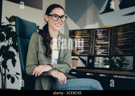 Young female programmer working in a modern office setting with monitors displaying code, showcasing technology skills Stock Photo