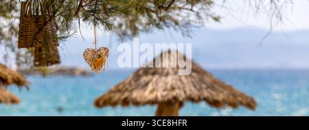 Romantic Tropical Beach Scene with Wicker Heart Decoration Hanging from Tree and Traditional Thatched Umbrella, panorama Stock Photo