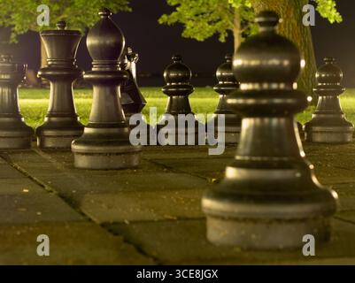 A striking low-angle shot of large, dark chess pieces on a tiled game board in a park at night. Stock Photo