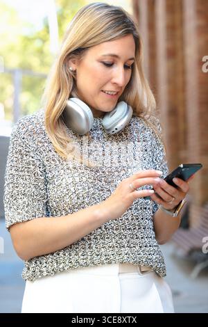 Young woman using smartphone on bench, headphones smiling outdoors Stock Photo