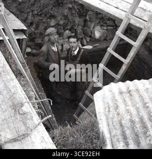 smoking cigarettes in garage 1950s, historical, two workmen having a break, smoking cigarettes, as they  stand by a wooden ladder in an underground pit, possibly a garage or  vehicle inspection pit, England, UK Stock Photo