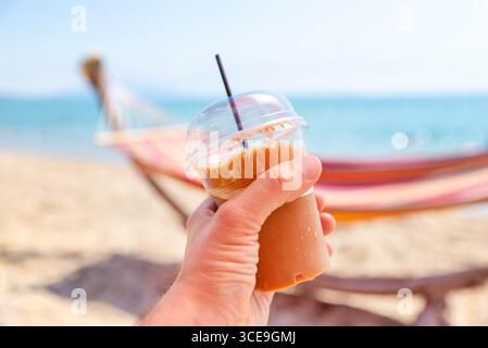 Hand holding iced coffee in a takeaway cup on a sunny beach, colorful hammock blurred in the background Stock Photo