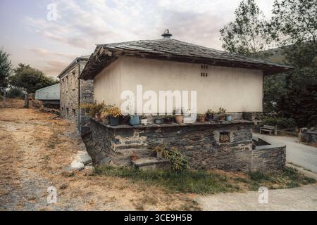 Traditional rustic stone and plaster house in Asturias, Spain, decorated with potted plants along the wall, surrounded by dry autumn grass and trees u Stock Photo