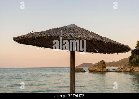 Straw beach umbrella at sunset with sea and rocky coastline in the background Stock Photo