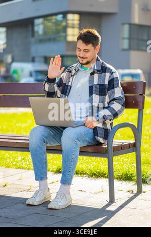 Mature businessman talking on video call using laptop computer while sitting on bench in city street. Male manager employer in casual clothes having an online conversation, remote business meeting. Stock Photo