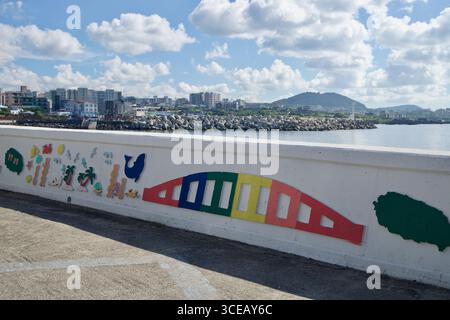 Colorful relief artwork decorates a harbor seawall in Iho‑dong, with tetrapods, apartment blocks, and a low volcanic hill rising across the quiet bay Stock Photo