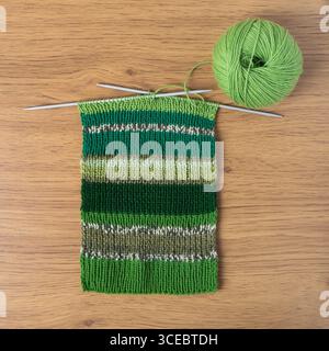 Green knitted wool on wooden background with knitting needles for knitting warm clothes and hobbies needlework. Knitting striped bright scarf made of yellow, green and light green threads. Stock Photo